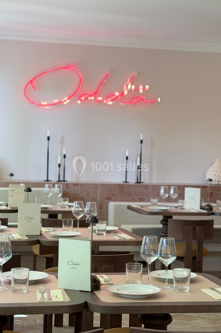 Salle de restaurant élégante avec tables dressées, verres, menus et enseigne lumineuse ’Odela’ au mur.