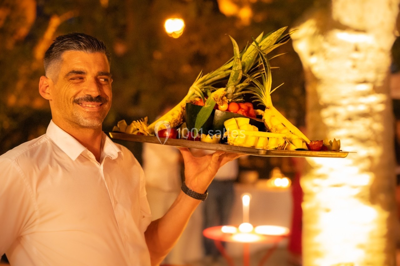 Un serveur souriant porte un plateau garni de fruits tropicaux, en extérieur, éclairé par des lumières chaudes.