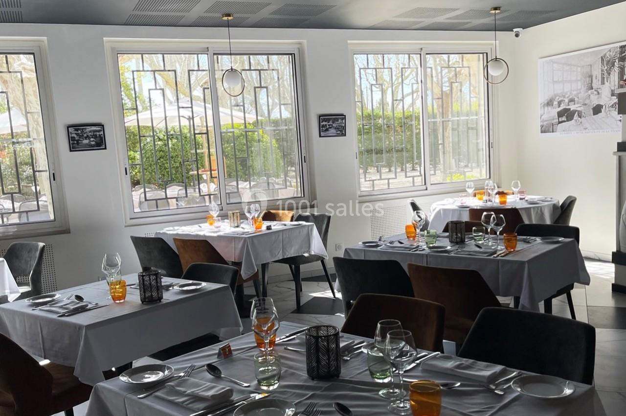 Salle de restaurant lumineuse avec tables dressées, nappes blanches, chaises variées et grandes fenêtres donnant sur l…