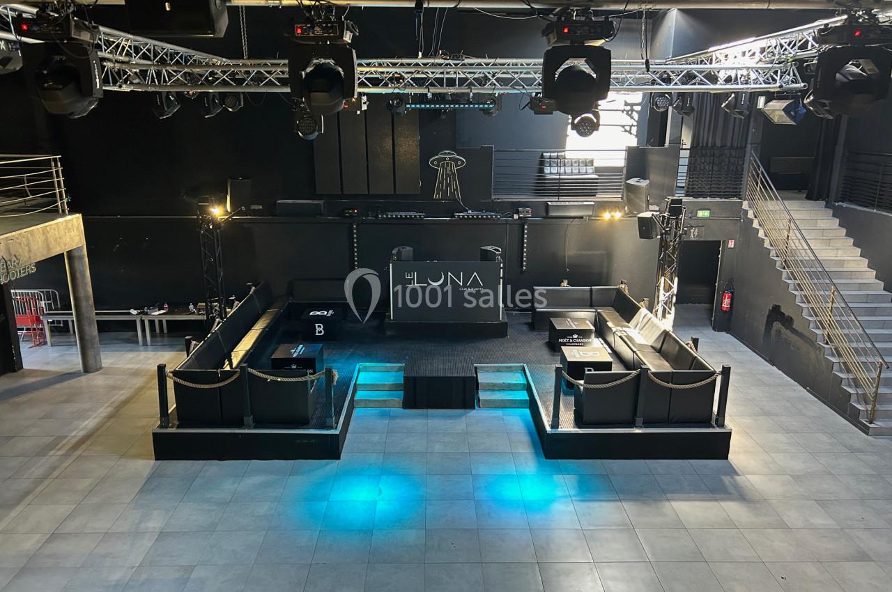 Salle de concert vide avec scène centrale, éclairages suspendus, estrades et escaliers sur les côtés.