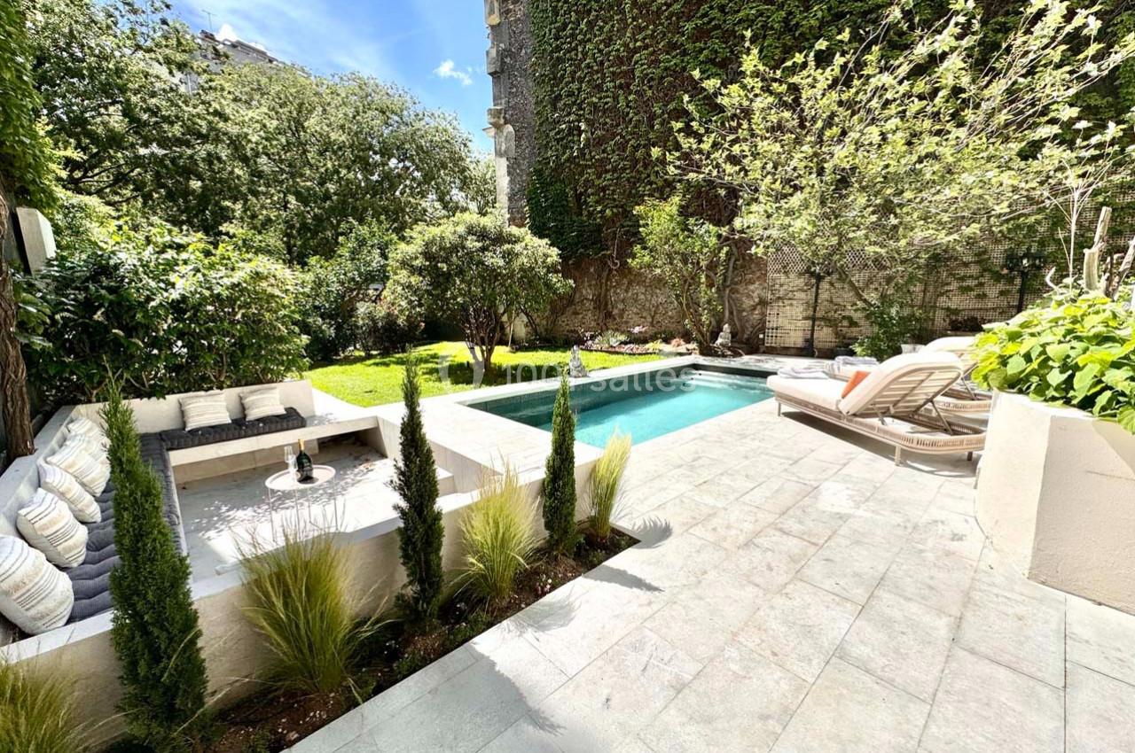 Jardin avec piscine, terrasse en pierre, coin salon avec coussins et végétation luxuriante sur un mur en arrière-plan.