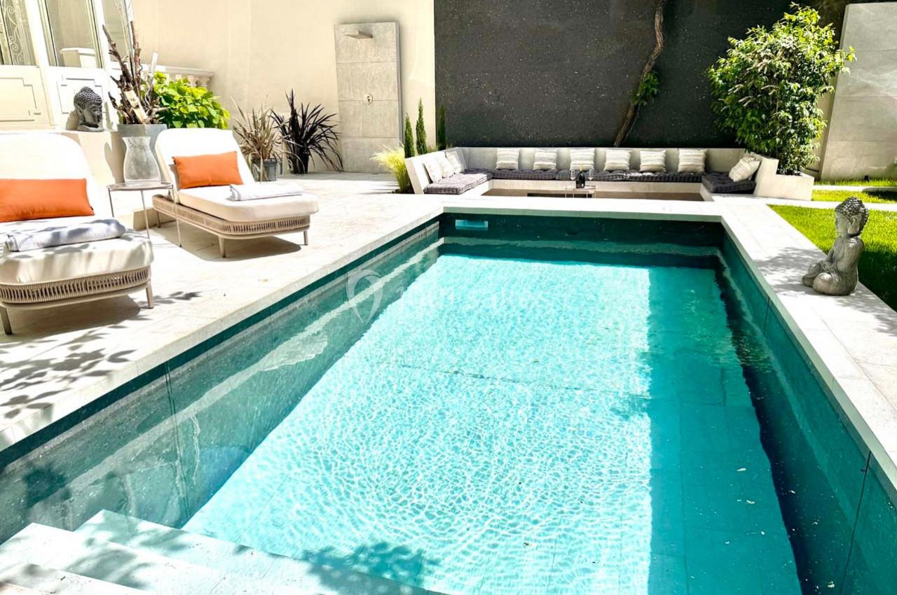 Piscine rectangulaire entourée de chaises longues, canapé d'extérieur et végétation dans une cour ensoleillée.