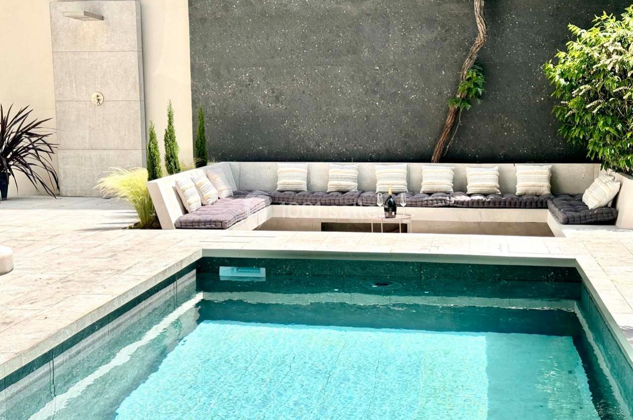 Piscine extérieure avec eau turquoise, terrasse en pierre, canapé d'angle avec coussins et mur végétalisé en arrière-plan.