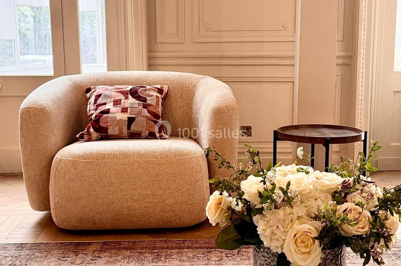 Fauteuil beige avec coussin décoratif, table d'appoint en bois et bouquet de roses blanches dans un intérieur lumineux.