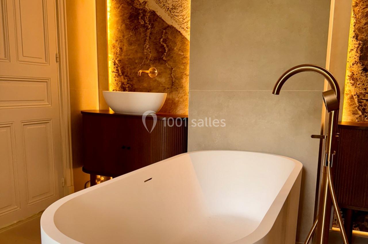 Salle de bain moderne avec baignoire îlot blanche, vasque ronde sur meuble en bois et éclairage chaleureux.