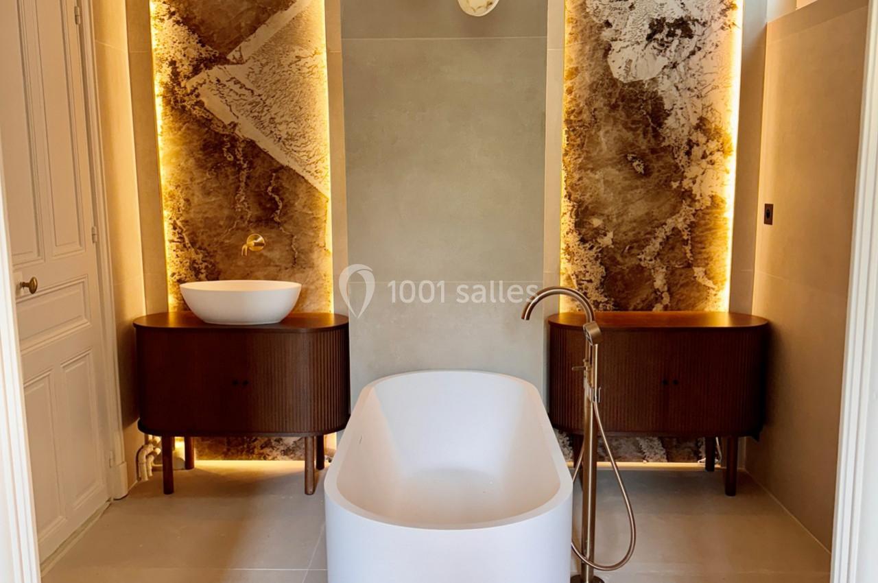 Salle de bain moderne avec baignoire centrale, deux vasques sur meubles en bois et éclairage mural chaleureux.