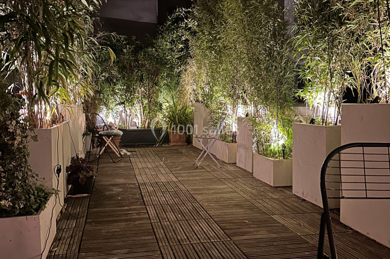Terrasse en bois éclairée la nuit, entourée de plantes en pots et équipée de chaises et d'une table.