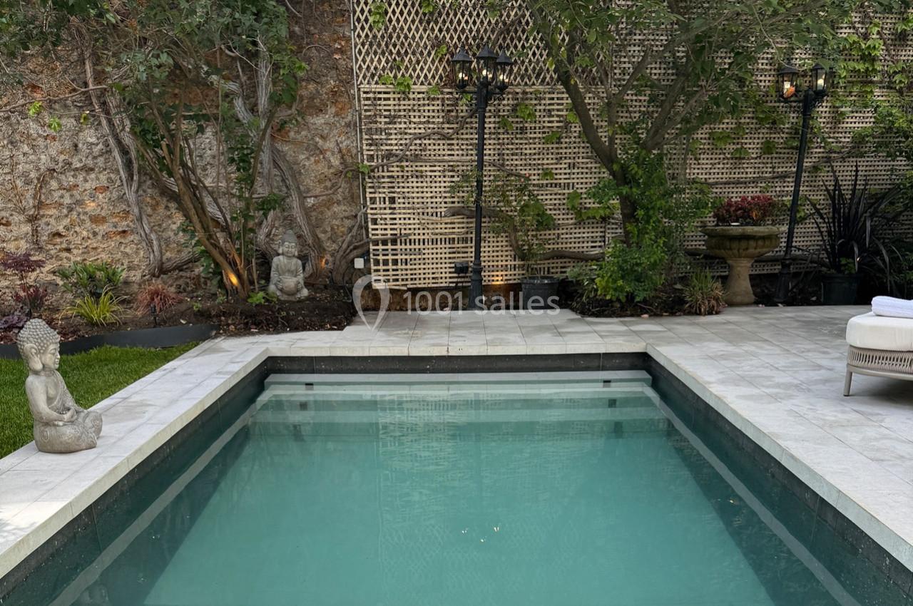 Piscine rectangulaire entourée de statues, végétation et éclairages, dans un jardin avec mur en pierre et treillis.