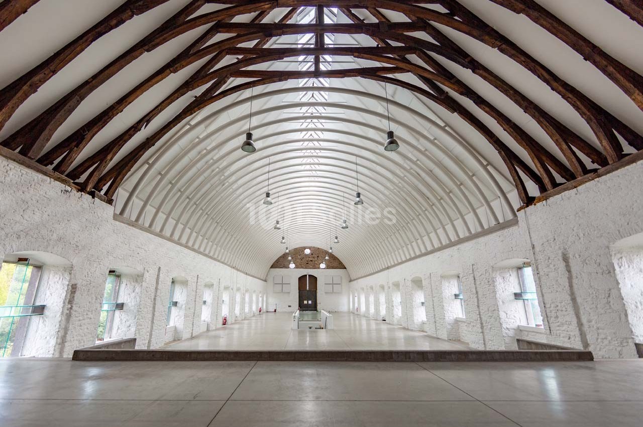 Grande salle voûtée avec poutres en bois apparentes, murs blancs en pierre et fenêtres alignées sur les côtés.