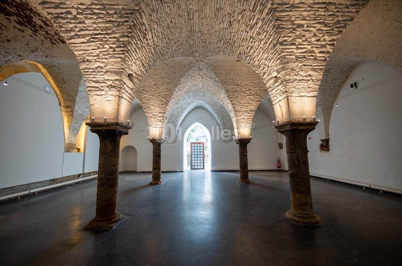 Salle voûtée en pierre avec colonnes centrales, éclairage doux et sol sombre, donnant sur une porte en arrière-plan.