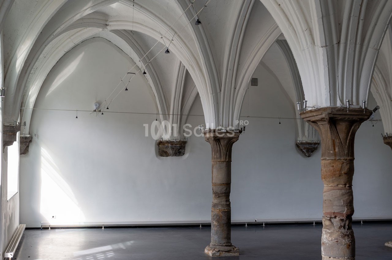 Salle vide avec des colonnes en pierre et des voûtes gothiques blanches sous un éclairage naturel.