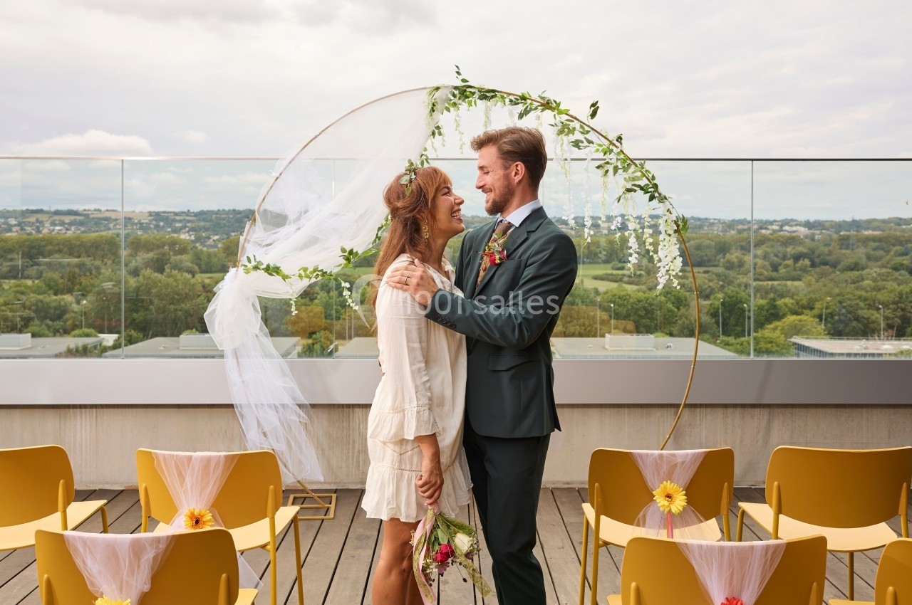 Un couple se tient sous une arche décorée de fleurs sur une terrasse avec vue sur un paysage verdoyant.