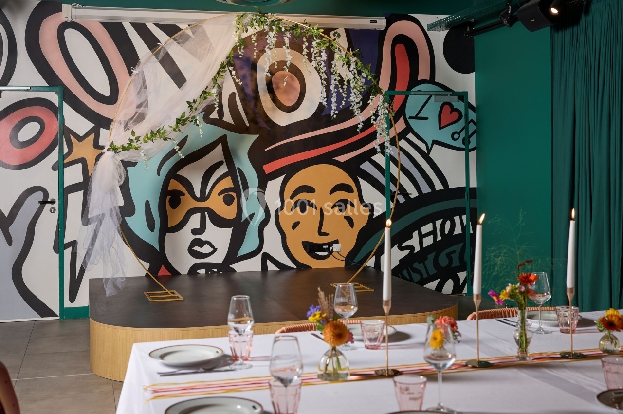 Décoration de table avec fleurs et bougies devant une arche ornée de feuillage et un mur coloré aux motifs artistiques.