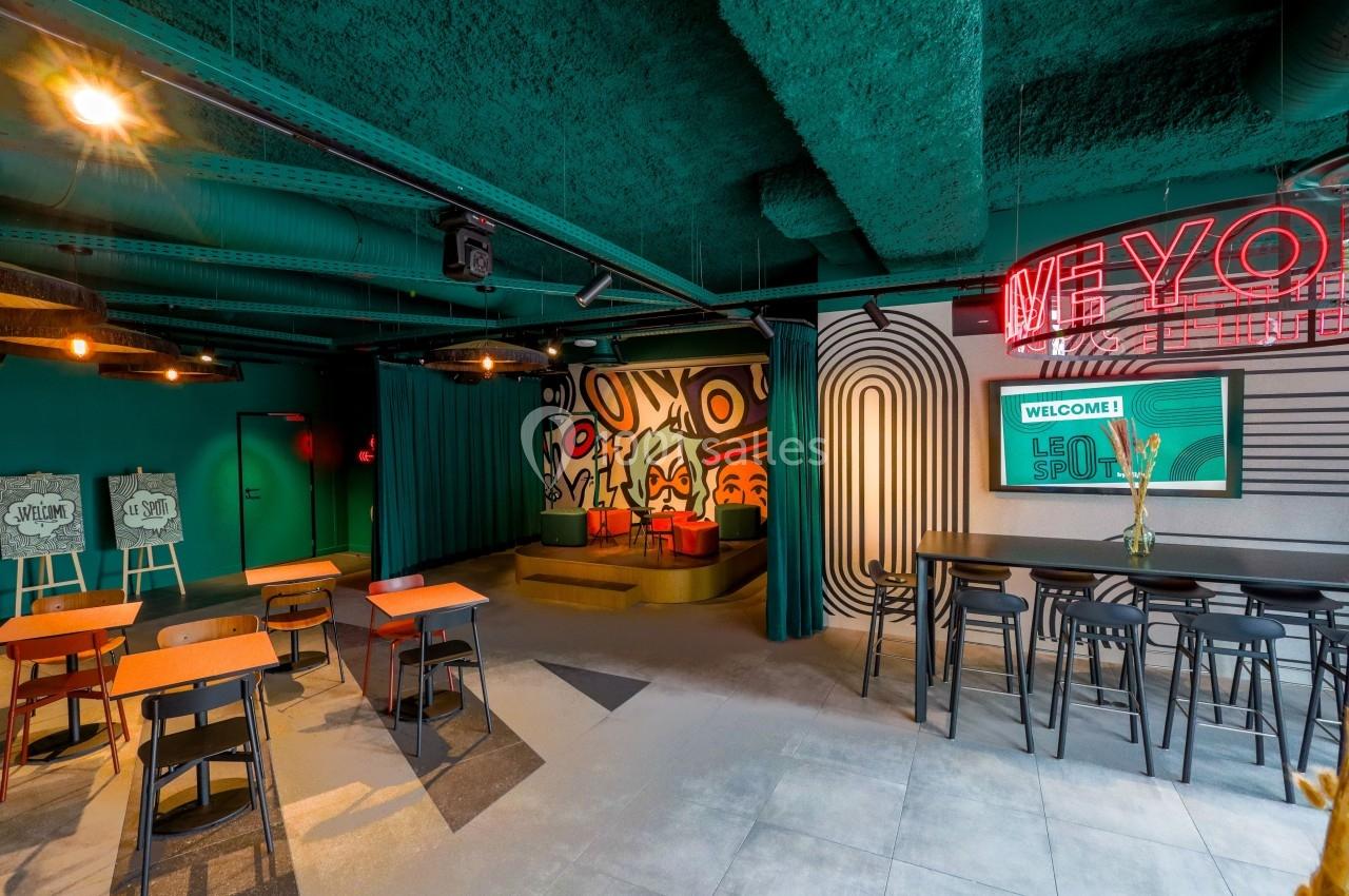 Espace intérieur moderne avec tables, chaises, scène décorée et enseignes lumineuses dans une ambiance colorée.