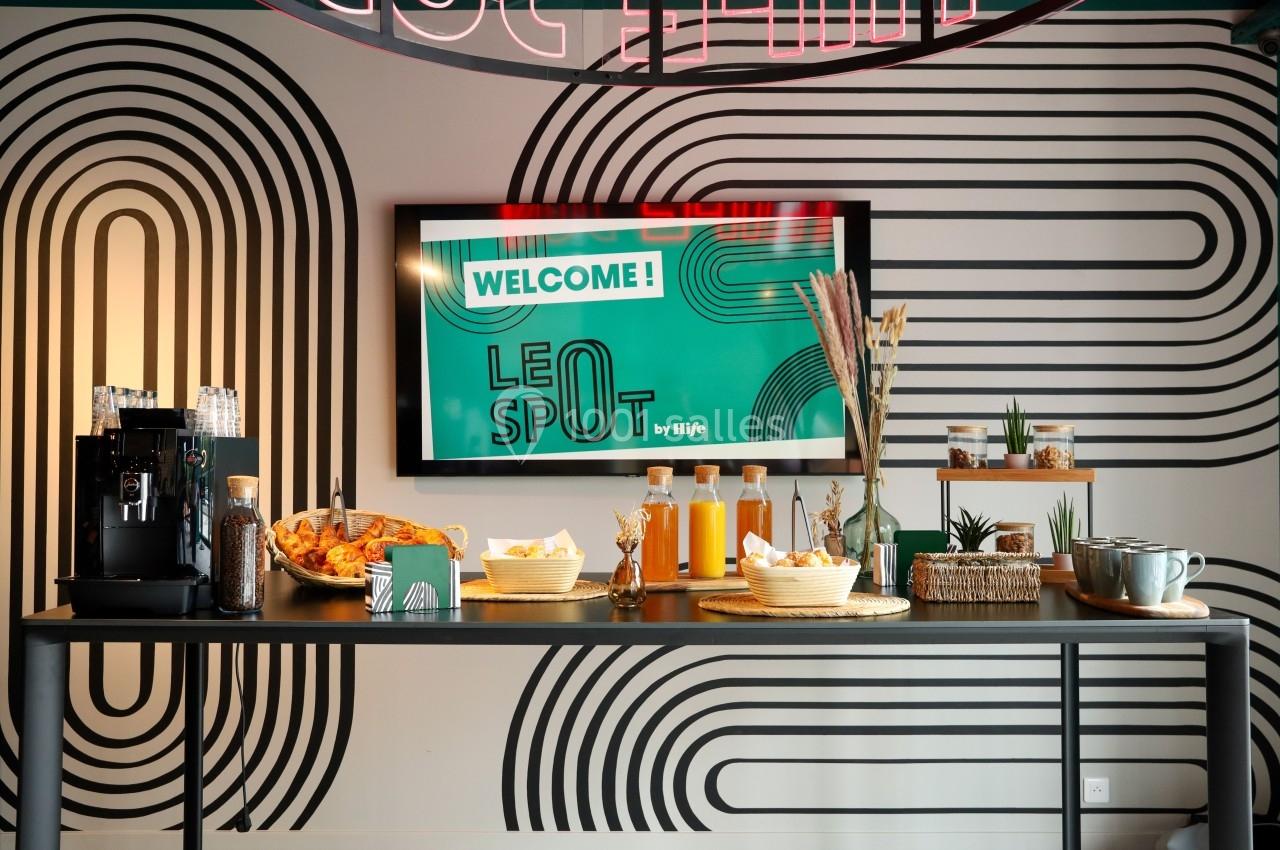 Table avec boissons, viennoiseries et décorations, devant un mur graphique et un écran affichant ’Welcome! Le Spot’.