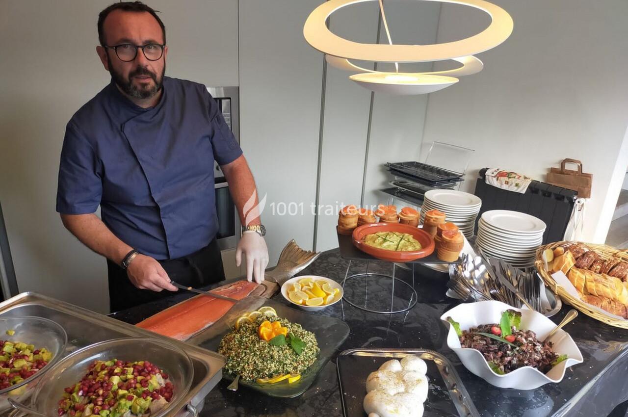 Un chef découpe du saumon fumé sur un buffet composé de plats variés, pains et desserts dans une cuisine moderne.