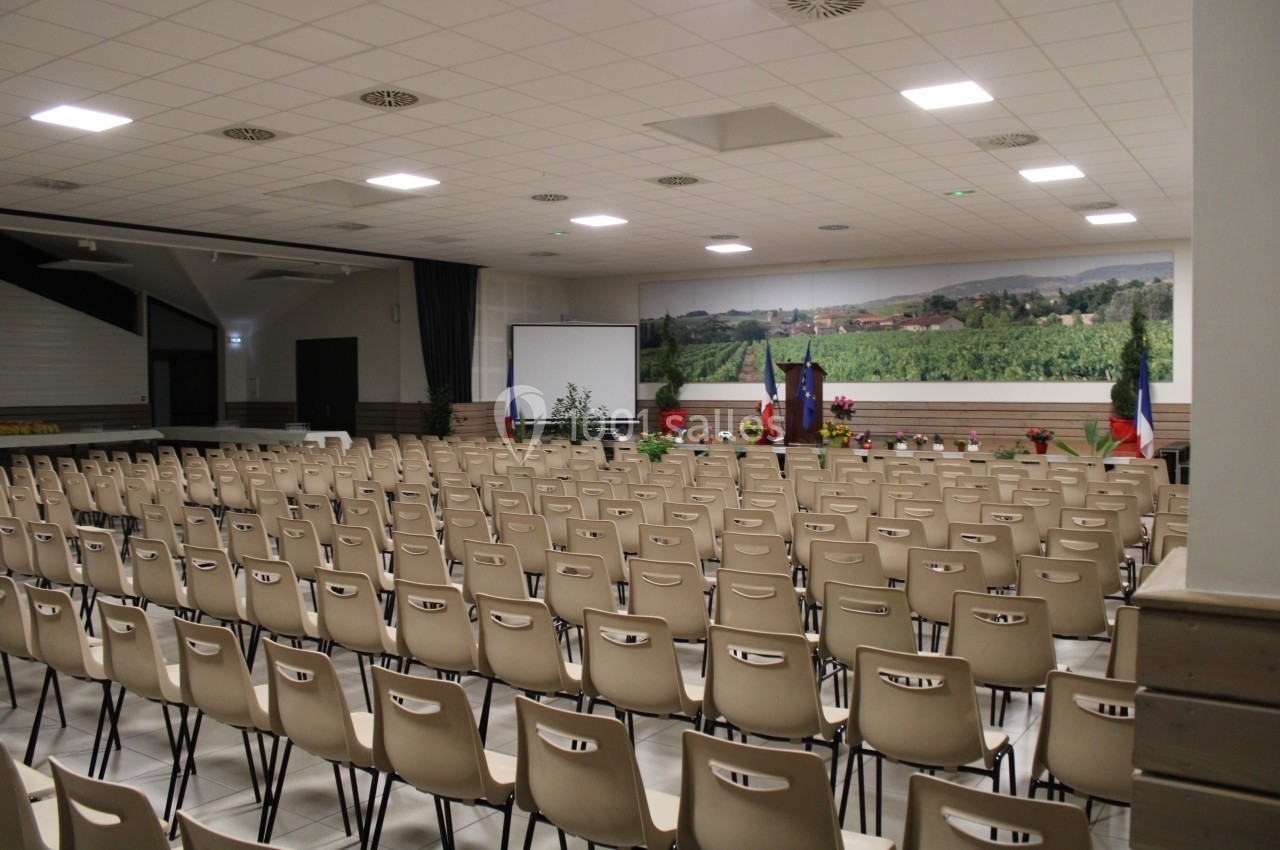 Salle de conférence vide avec des rangées de chaises beiges, un écran de projection et des drapeaux en fond.