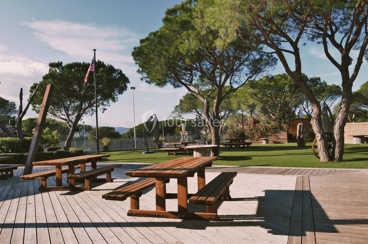 Tables de pique-nique en bois sur une terrasse ensoleillée, entourées d'arbres et d'une aire de jeux en arrière-plan.