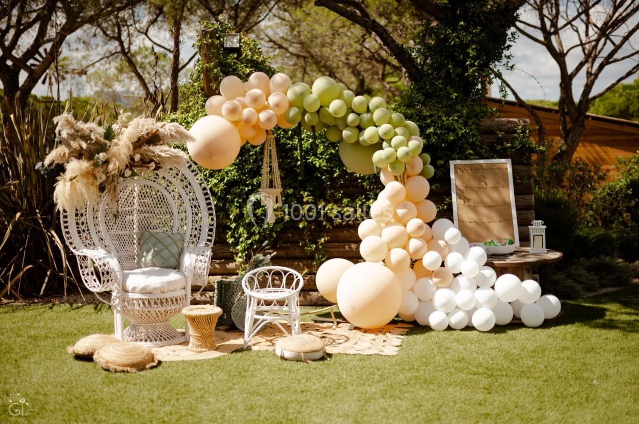 Décoration extérieure avec fauteuil en rotin, arche de ballons pastel et éléments naturels sur pelouse.