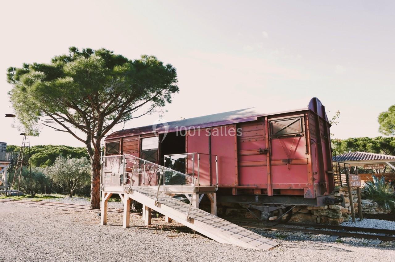 Wagon rouge aménagé avec une rampe d'accès en bois, situé près d'un arbre et entouré de végétation.