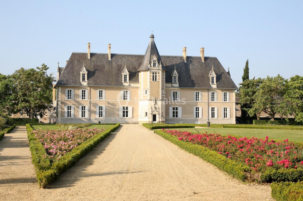Façade d'un château entouré de jardins fleuris et d'une allée centrale en gravier, sous un ciel dégagé.
