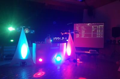 Un DJ mixe sur une scène éclairée par des lumières colorées dans une salle sombre avec un écran projeté.