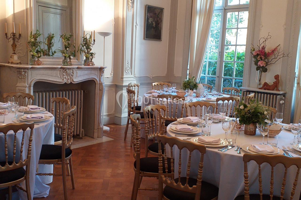 Salle élégante avec tables rondes dressées, nappes blanches, chaises en bois doré et décorations florales.