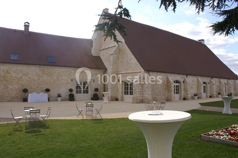 Bâtiment en pierre avec toit en pente, tables et chaises disposées sur une terrasse devant une pelouse.