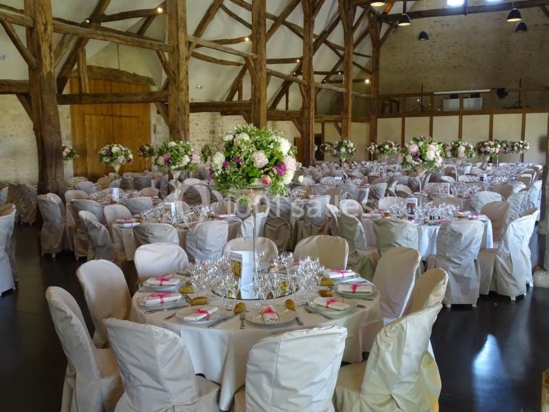 Salle de réception décorée pour un mariage, avec tables rondes dressées, chaises couvertes et arrangements floraux.