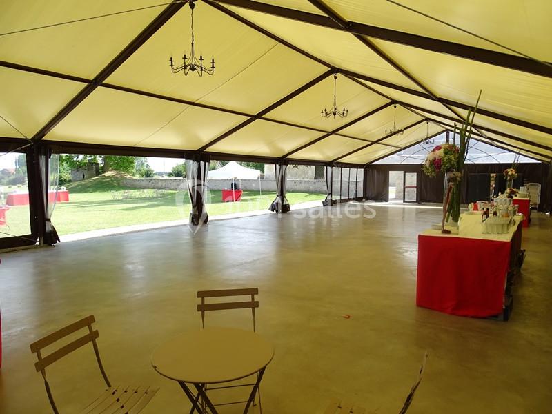 Intérieur d'une grande tente événementielle avec sol en béton, tables, chaises et vue sur un espace extérieur verdoyant.