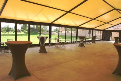 Bâtiment en pierre avec toit en pente, tables et chaises disposées sur une terrasse et une pelouse au premier plan.