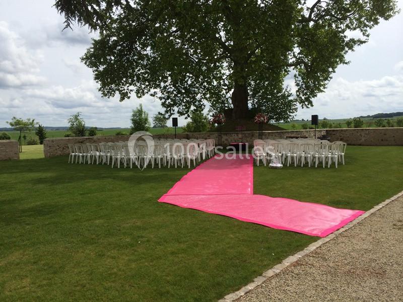 Allée rose menant à des chaises blanches disposées en extérieur sous un grand arbre, pour une cérémonie.