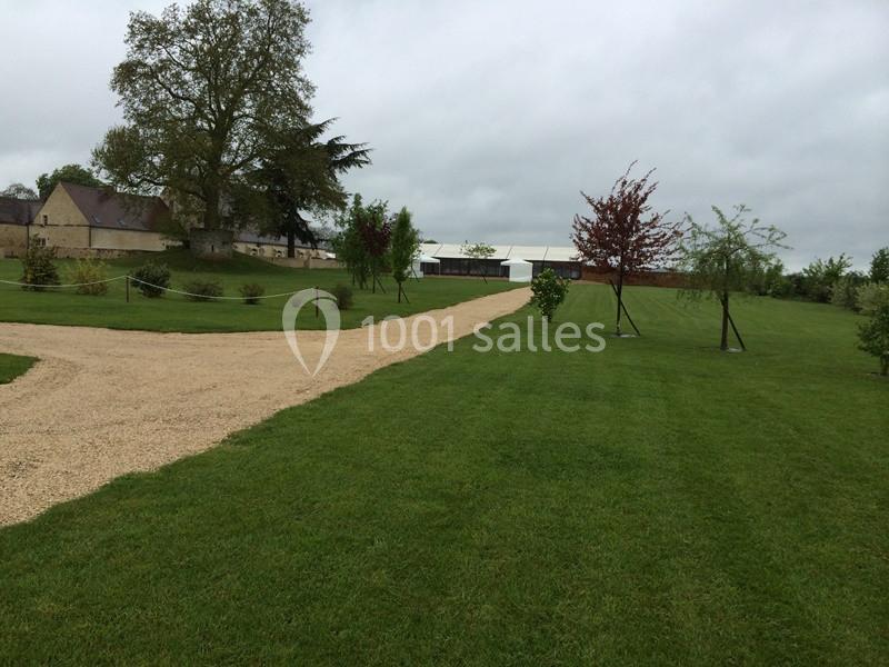 Allée en gravier bordée de pelouses et d'arbres, menant à une grande tente blanche dans un paysage rural.