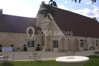 Bâtiment en pierre avec toit en pente, tables et chaises disposées sur une terrasse et une pelouse au premier plan.