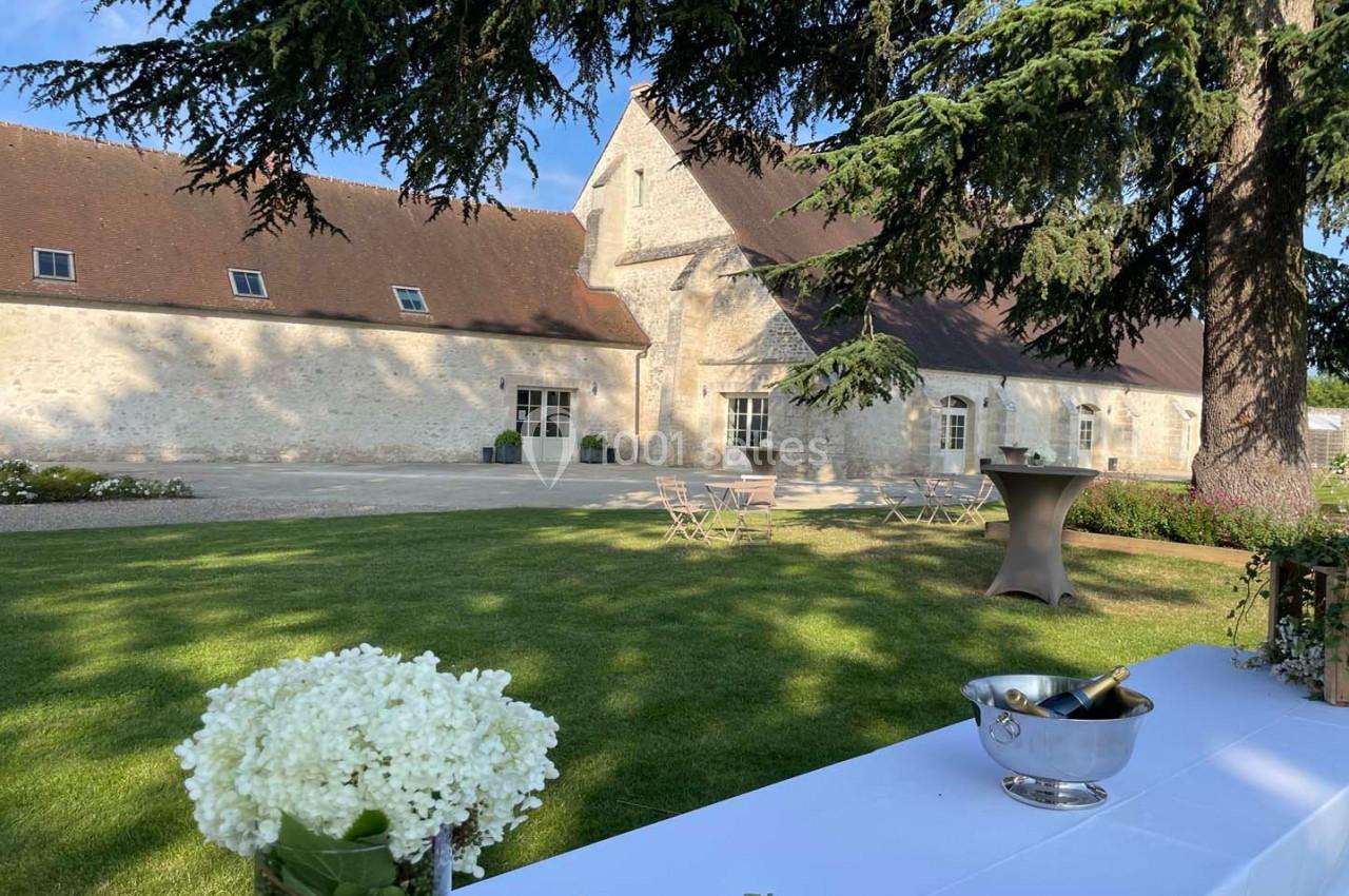 Cour d'une bâtisse en pierre avec pelouse, tables de jardin, et une table décorée de fleurs blanches au premier plan.