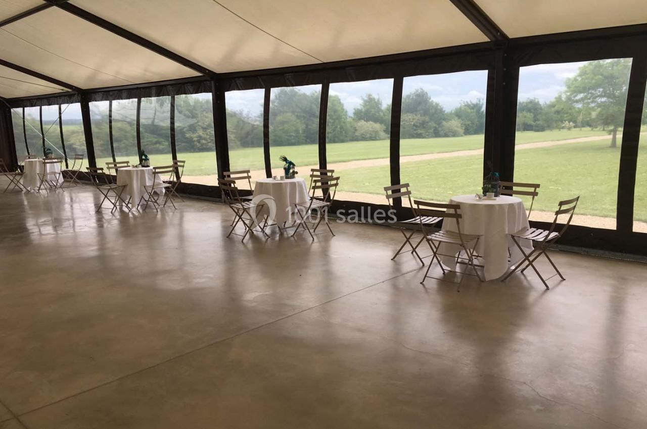 Salle avec de grandes baies vitrées donnant sur un espace vert, tables rondes dressées avec chaises en bois pliantes.