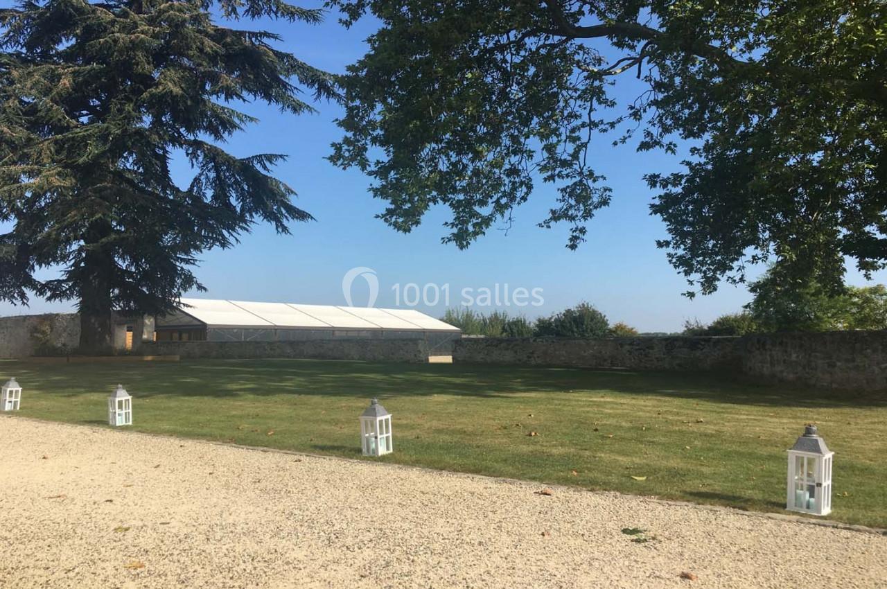 Pelouse avec lanternes blanches alignées, grand arbre, mur en pierre et tente blanche en arrière-plan.