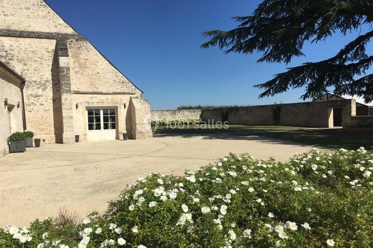 Cour ensoleillée avec façade en pierre, parterre de fleurs blanches au premier plan et mur en pierre en arrière-plan.