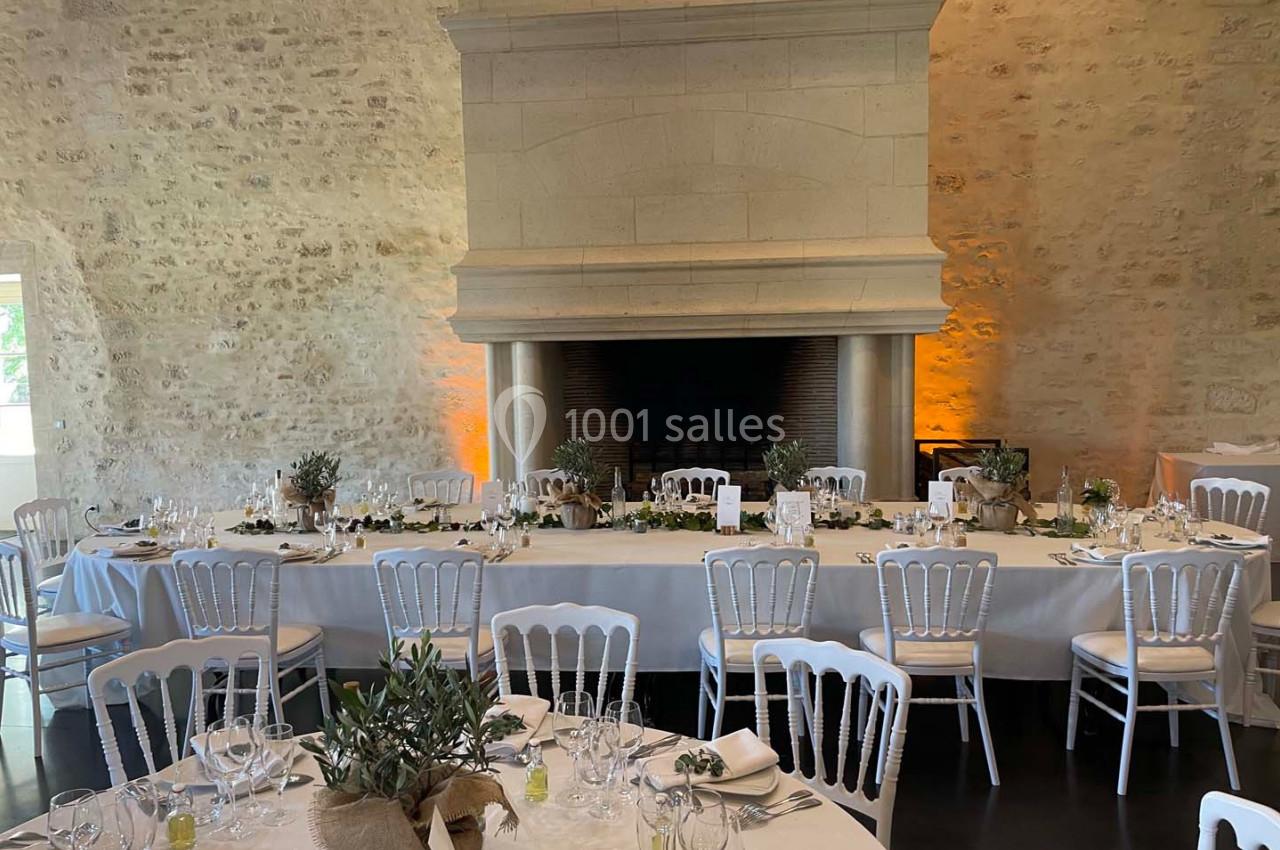 Salle de réception avec tables dressées, nappes blanches, chaises en bois et décoration végétale devant une grande cheminée.