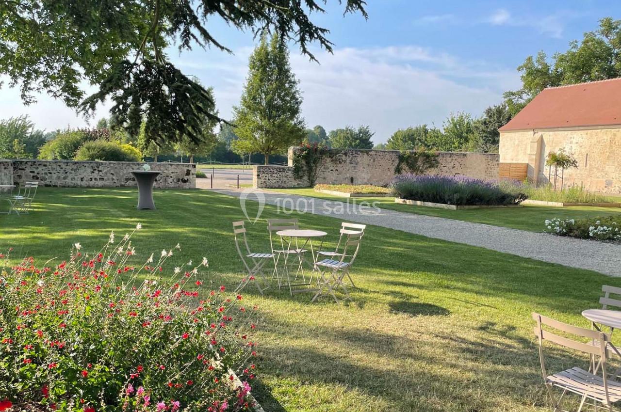 Jardin avec pelouse, tables et chaises en métal, bordé de fleurs, arbres et murs en pierre sous un ciel dégagé.