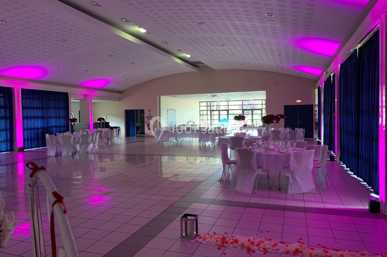 Salle de réception décorée avec des tables dressées, éclairage rose et rideaux bleus, prête pour un événement.