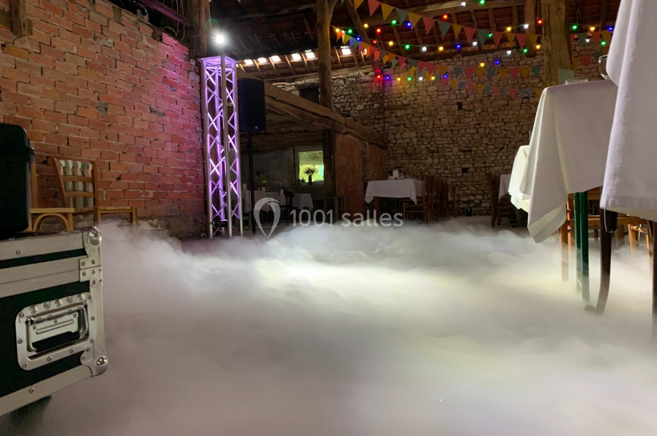 Salle en briques décorée avec des guirlandes lumineuses, tables dressées et sol couvert de fumée artificielle.
