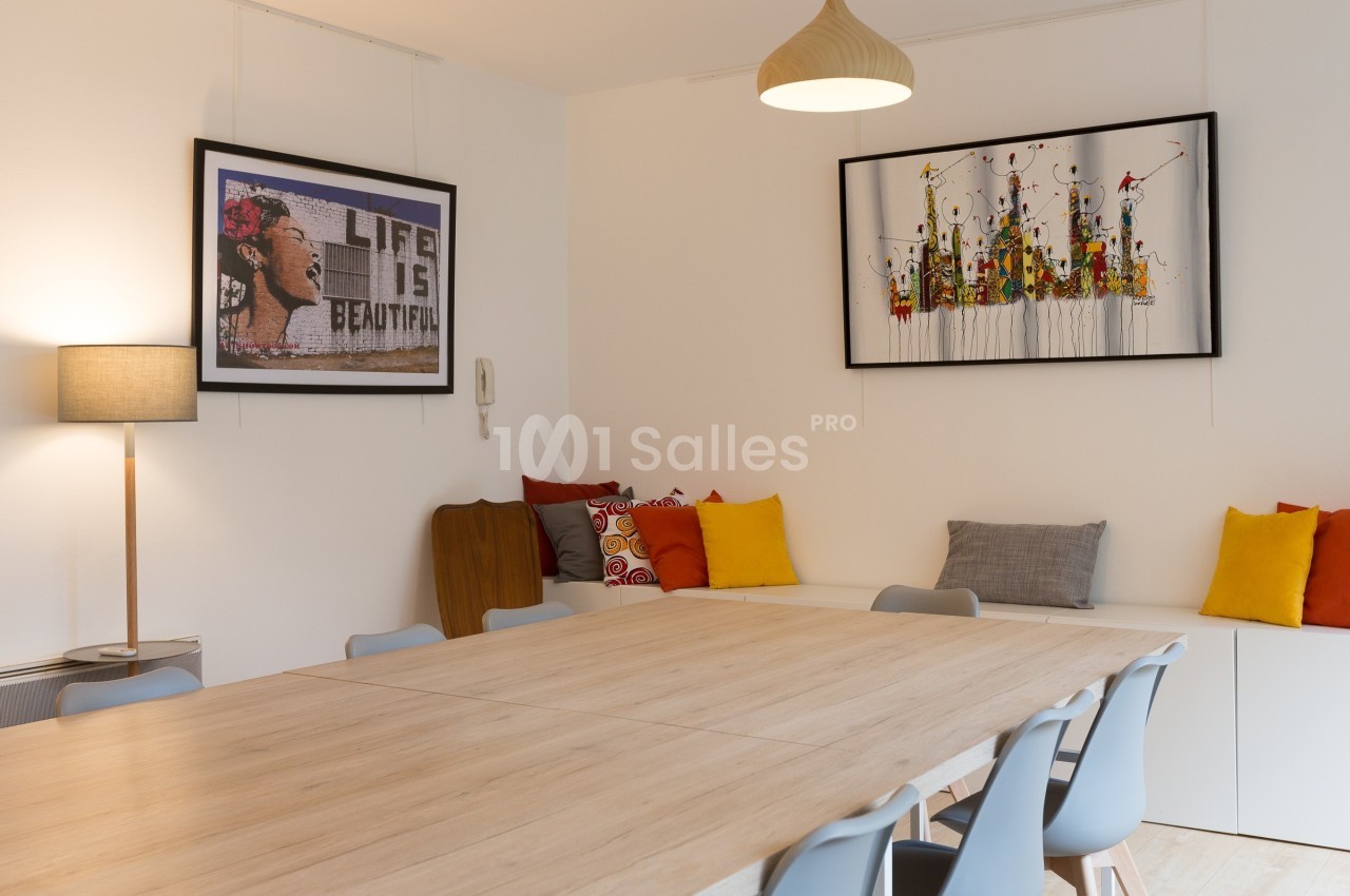 Salle à manger lumineuse avec grande table en bois, chaises grises, coussins colorés et tableaux décoratifs au mur.