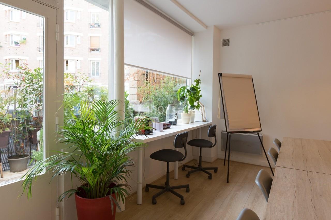 Salle lumineuse avec une grande fenêtre, des plantes, un bureau avec chaises, et un tableau de conférence.