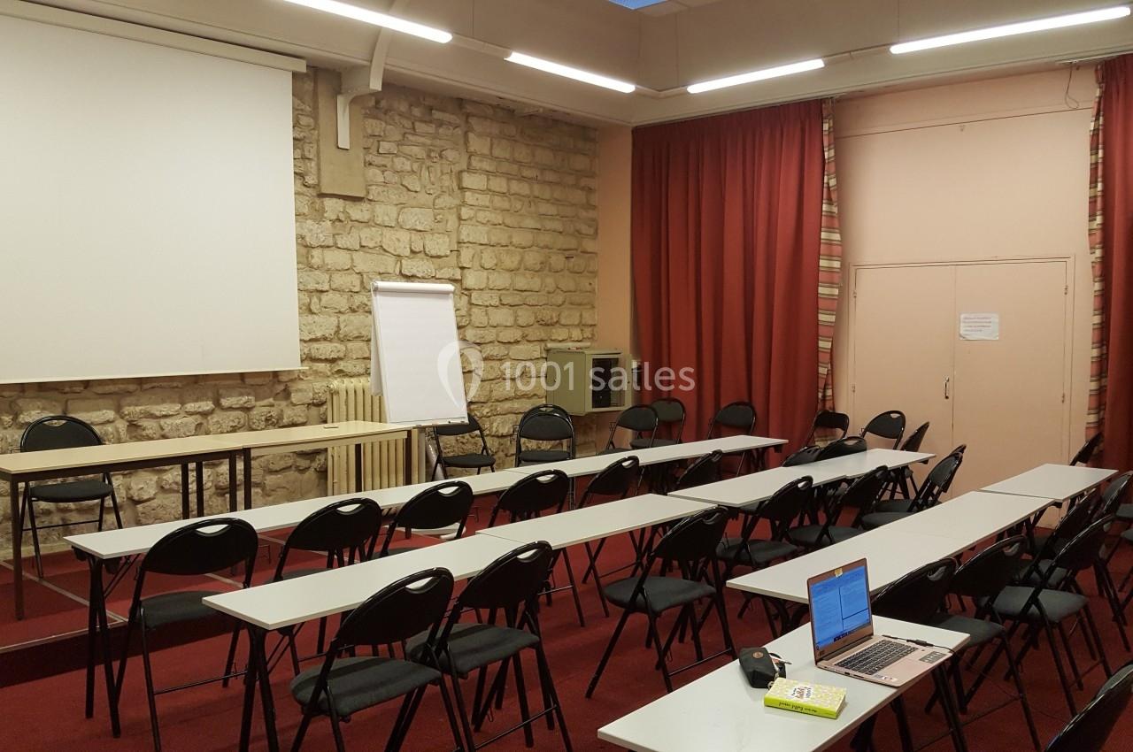 Salle de réunion avec tables, chaises alignées, tableau blanc, écran de projection et rideaux rouges.
