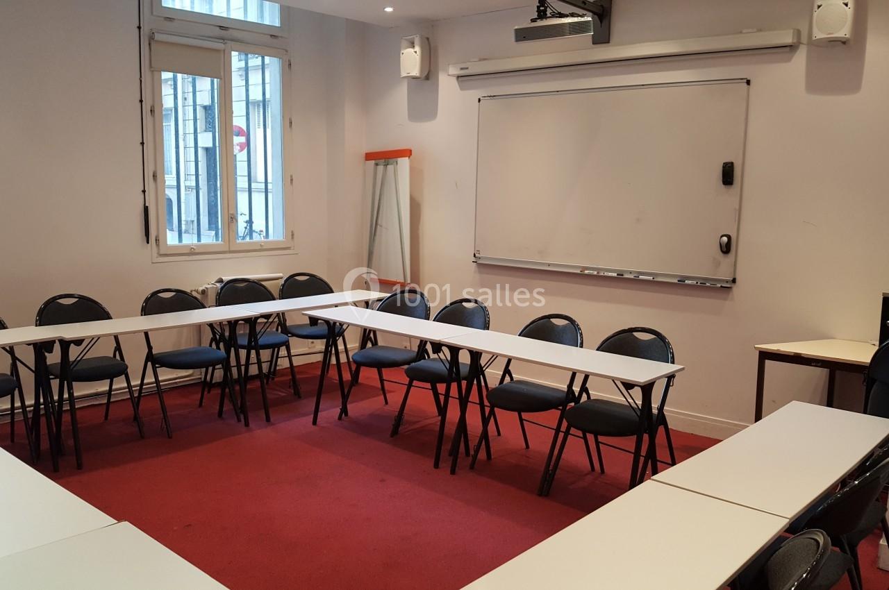 Salle de réunion avec tables disposées en U, chaises noires, tableau blanc et fenêtre donnant sur l'extérieur.