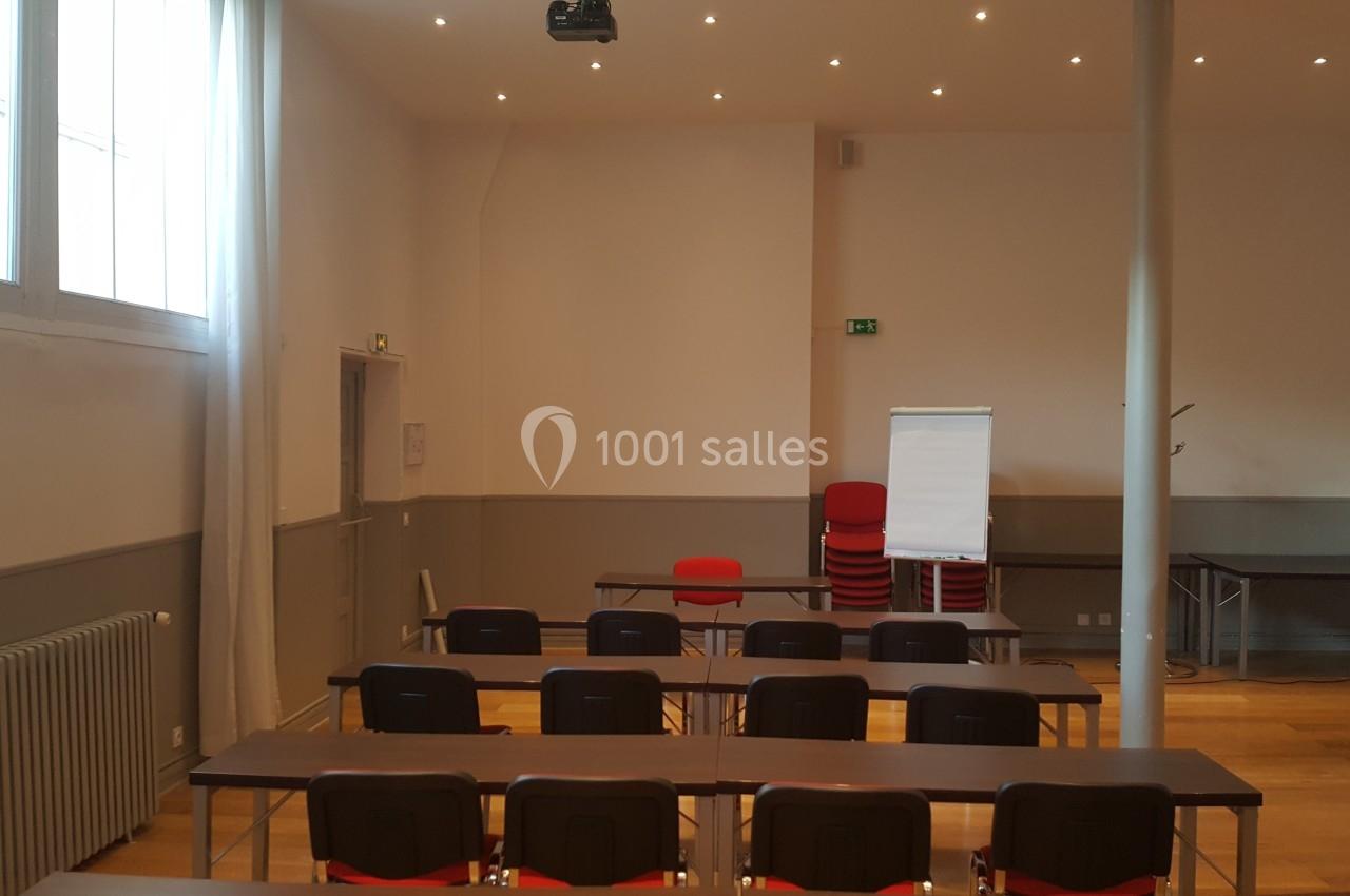 Salle de réunion lumineuse avec des tables, des chaises alignées, un paperboard et un projecteur au plafond.