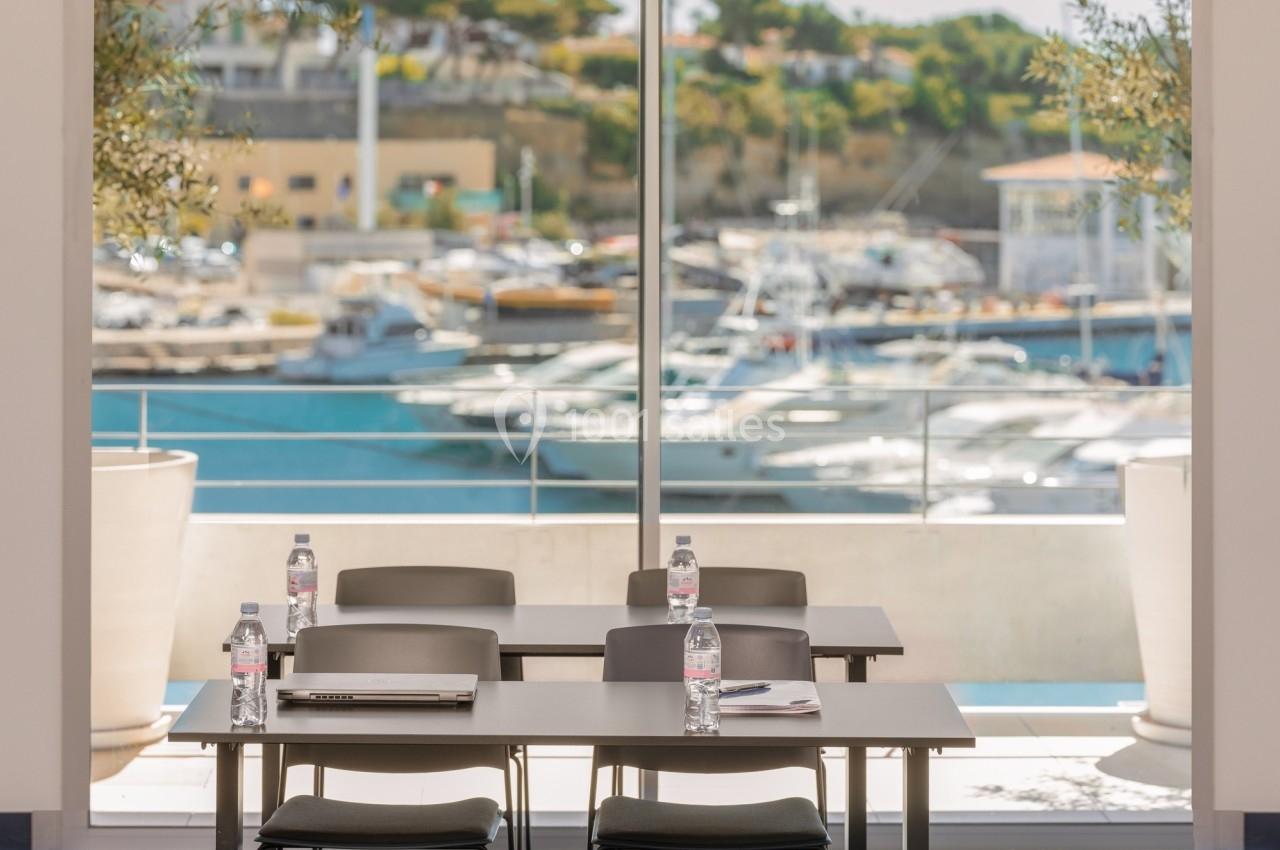 Salle de réunion lumineuse avec vue sur un port de plaisance, équipée de tables, chaises et bouteilles d'eau.
