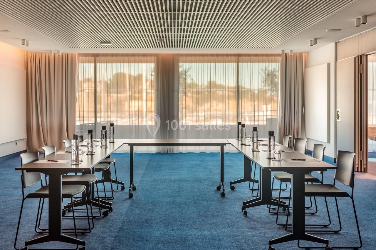 Salle de réunion lumineuse avec tables en U, chaises modernes et grandes baies vitrées donnant sur l'extérieur.