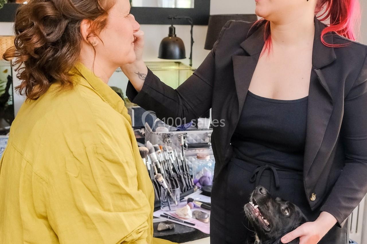 Une maquilleuse applique du maquillage sur une femme assise, un chien noir et blanc posé sur ses genoux.